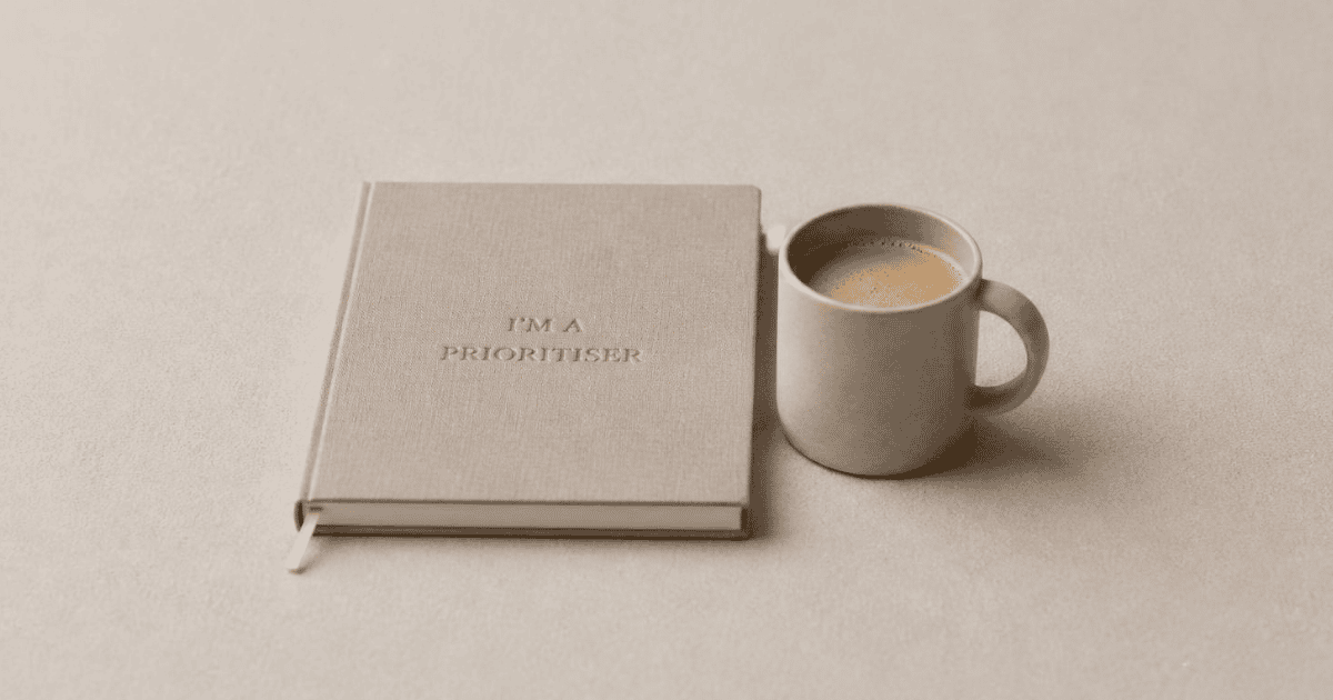 Notebook embossed with I'm a Prioritiser next to a cup of coffee on a beige surface